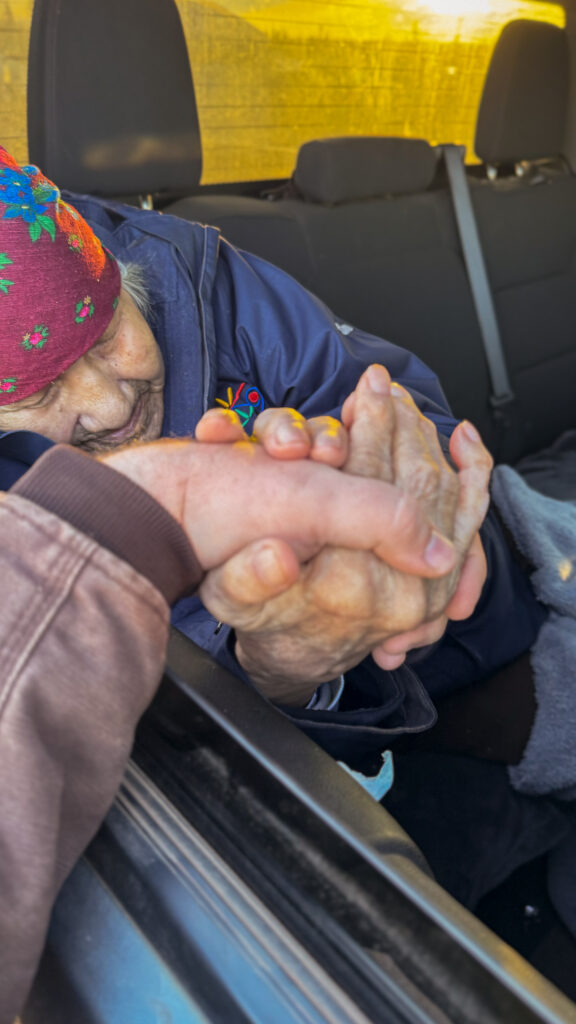  The same elderly woman, still holding onto the other person’s hand, appears deeply connected and emotionally engaged in the moment. Her grip is strong as she leans out from the car window. The background shows the interior of the vehicle, with warm sunlight reflecting inside.
