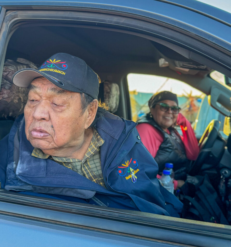 An elderly man sits in the passenger seat of a car, wearing a baseball cap with "Kawawachikamach" embroidered on it. His expression is calm, looking out the window. In the driver's seat is a woman smiling, also wearing a cap and sunglasses. They are dressed warmly, and the scene appears to capture a relaxed moment between the two.