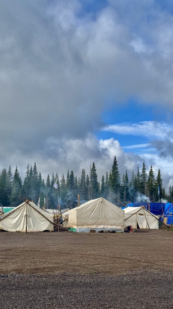 A row of canvas tents set up in a campsite, with smoke rising from campfires. The tents are surrounded by a forest of tall evergreen trees. Above the tents, the sky begins to clear, with bright blue patches breaking through the lingering storm clouds. The ground is wet from recent rain, but the sun is starting to shine, bringing a calm after the storm.