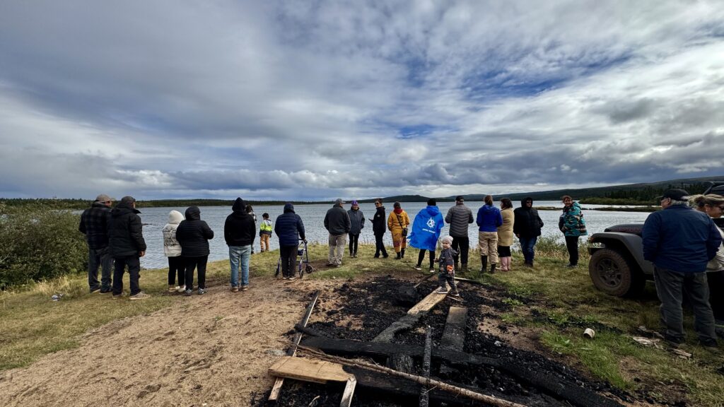 A group of people stand together on the shore of a lake, bundled up in warm jackets and coats. The cloudy sky looms above, and the lake reflects the gray light of the day. In the foreground, a burned pile of wood is visible on the ground.