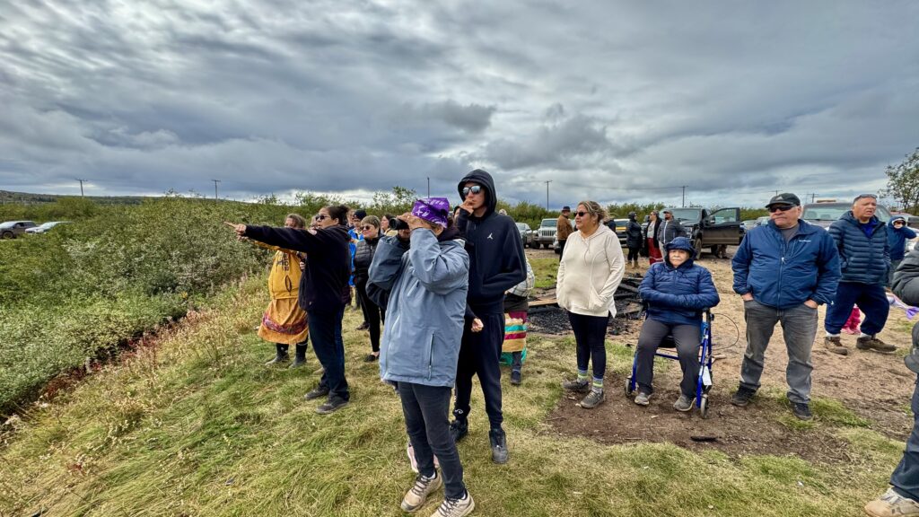 A group of people, some pointing and others looking on, stand by the lake. The sky is overcast, and a colorful variety of jackets and coats brighten the scene.