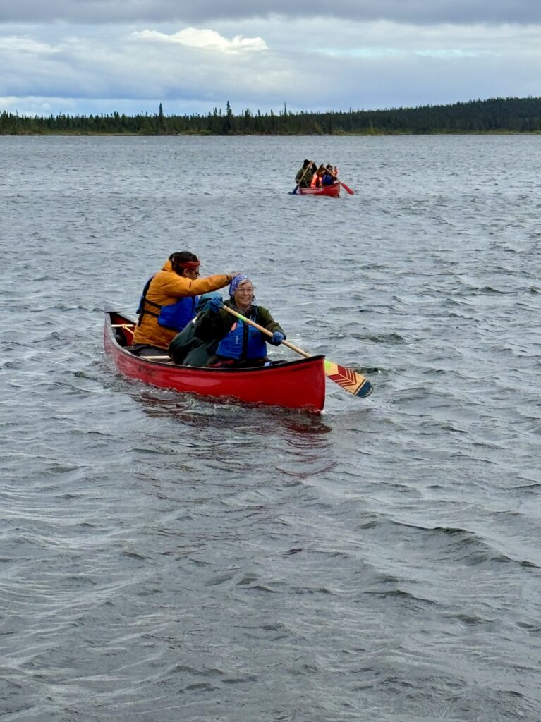 Two canoes, each with two paddlers, move across the water on a cloudy day. The water ripples in the wind, and the paddlers appear determined as they make their way towards the shore.