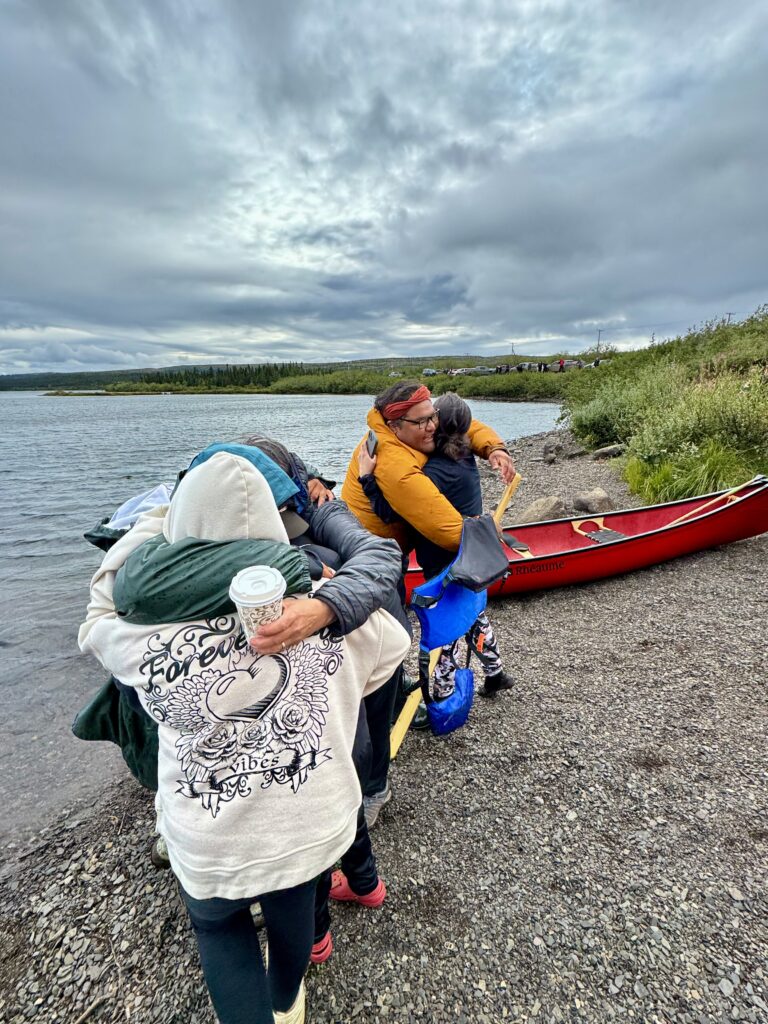 A woman in a rain jacket and boots steps onto the shore, greeting a young boy who runs toward her. A man in an orange jacket and life vest stands by a red canoe in the background. The lake's edge is rocky, with the water just a few feet away.