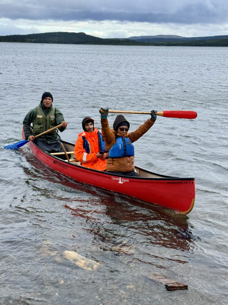 Three people in a red canoe, paddling on a lake. One person is raising their paddle in celebration while another looks at the camera. The backdrop features a cloudy sky and a forested shoreline.