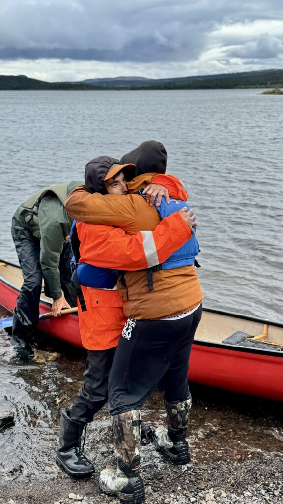 Two individuals embracing joyfully by the shore, with one man in a bright orange jacket and the other in a green jacket and hat. They are next to a red canoe, on a cloudy day with a lake in the background.