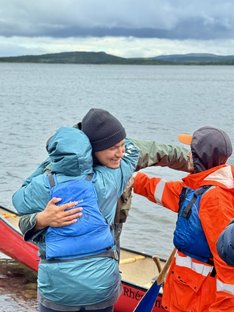 Two people hugging, one in a green jacket and the other in an orange jacket, beside a red canoe on a rocky shore. They are smiling and looking at each other with a lake and cloudy skies in the background.