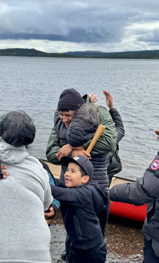 A festive scene beside a red canoe with several people embracing and celebrating. An adult is hugging a child, while others around them appear joyful. The setting includes a cloudy sky and a lake in the background.