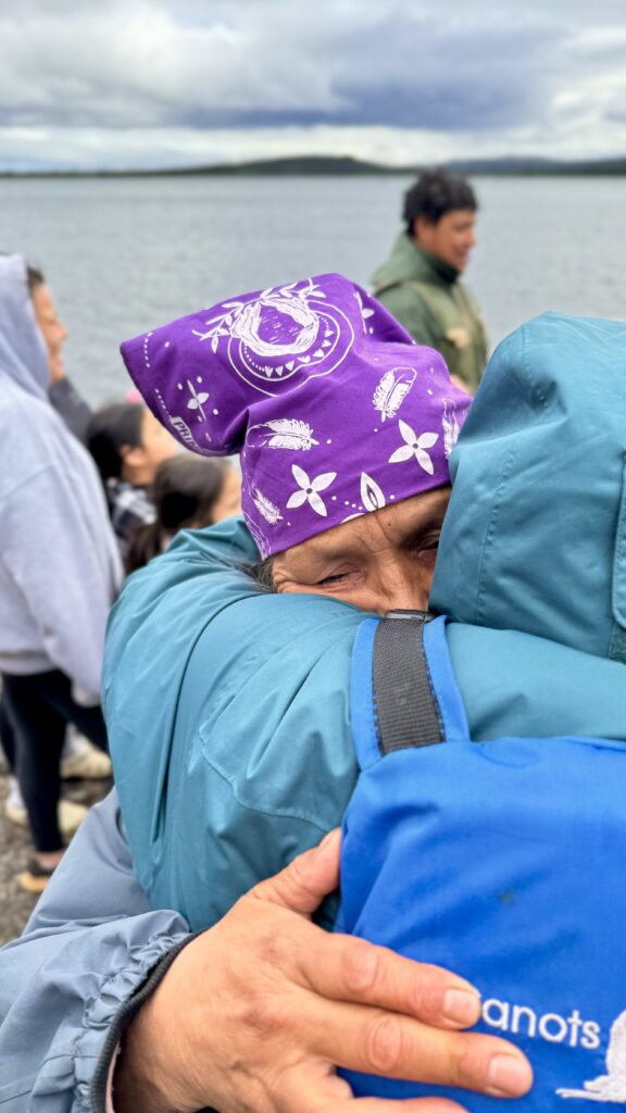 An emotional close-up of two individuals embracing by the lake. The person in a purple bandana and blue jacket closes their eyes in the hug, with other people standing near the water in the background.