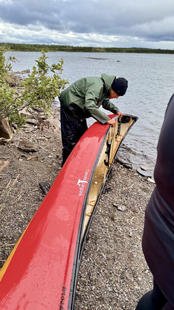 A person in a green rain jacket and black beanie carefully handling a red canoe on the shore of a lake. The canoe is positioned upside down on the rocky beach, and the surroundings include bushes and a cloudy sky.