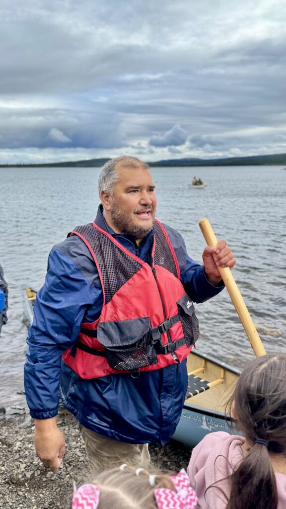 A man in a blue jacket with a red life vest holds a paddle and smiles while standing beside a lake. A canoe and small group can be seen in the background on the water, while children stand nearby on the shore.