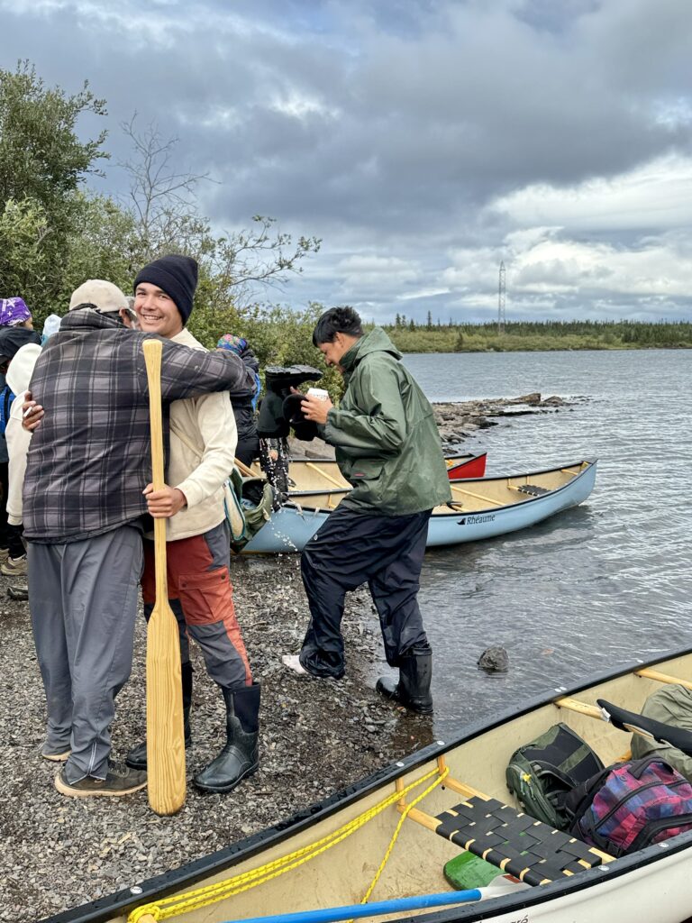 A group of people gathered at the lake's edge, including two individuals hugging with one holding a wooden paddle. Canoes rest on the shore as others engage in conversation and sip drinks. The sky is cloudy, with trees and water in the background.