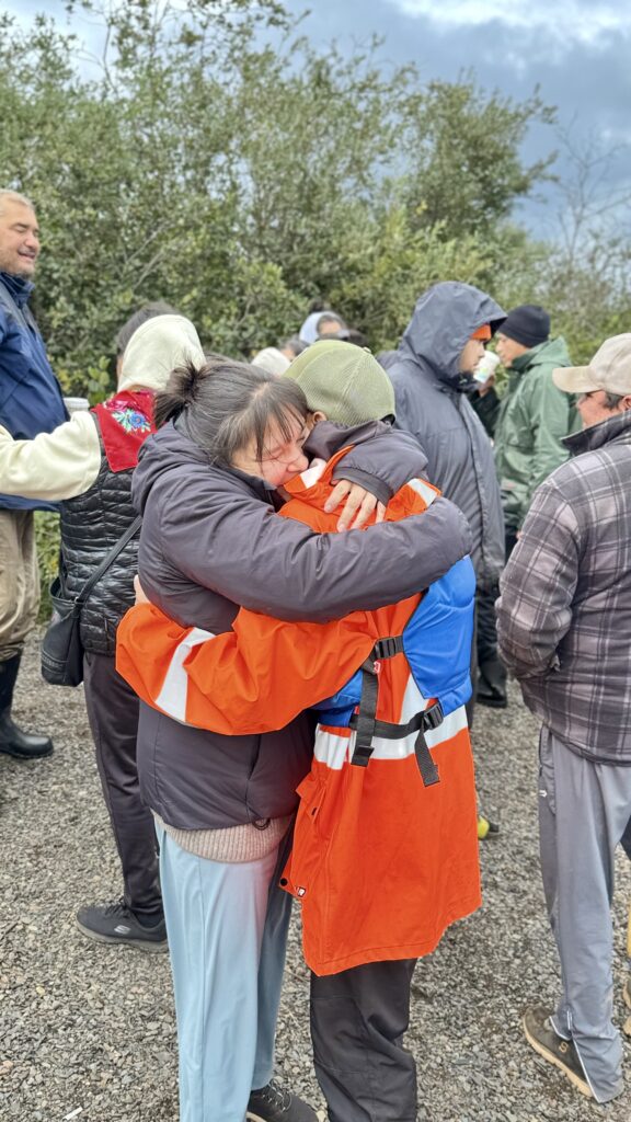 Two people share a warm embrace, one in a bright orange jacket and the other in a black coat. Others gather nearby, with trees and cloudy skies in the background as the community celebrates by the lakeshore.