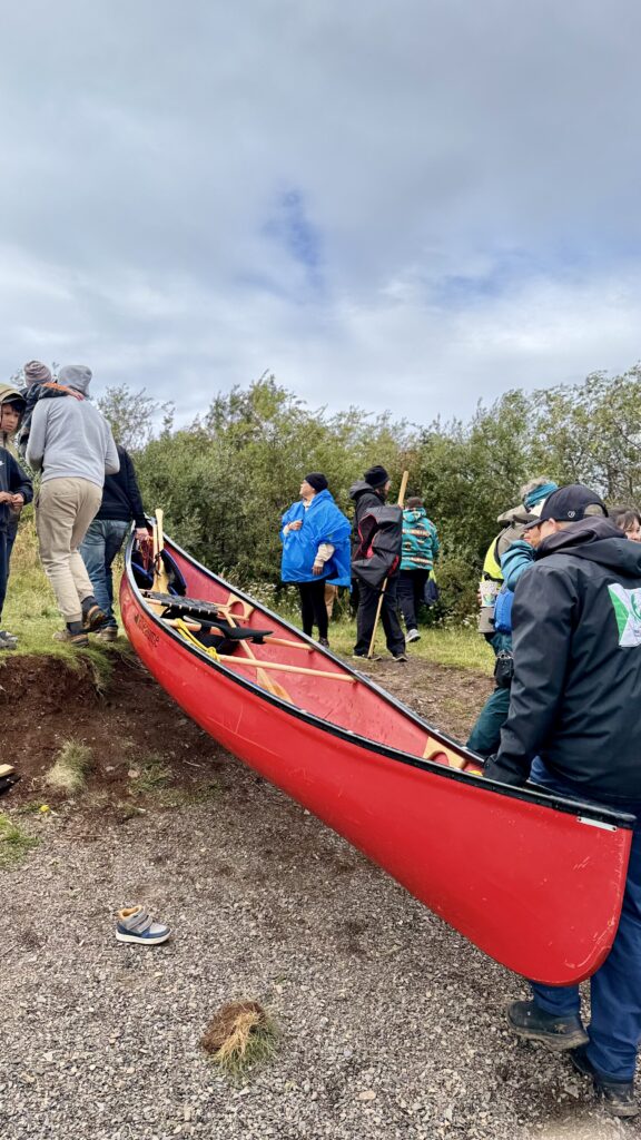 Several people work together to carry a large red canoe up a slight incline, while others gather nearby in small groups. The scene is set by the lake with a cloudy sky, and the group wears a mix of outdoor gear for the occasion.