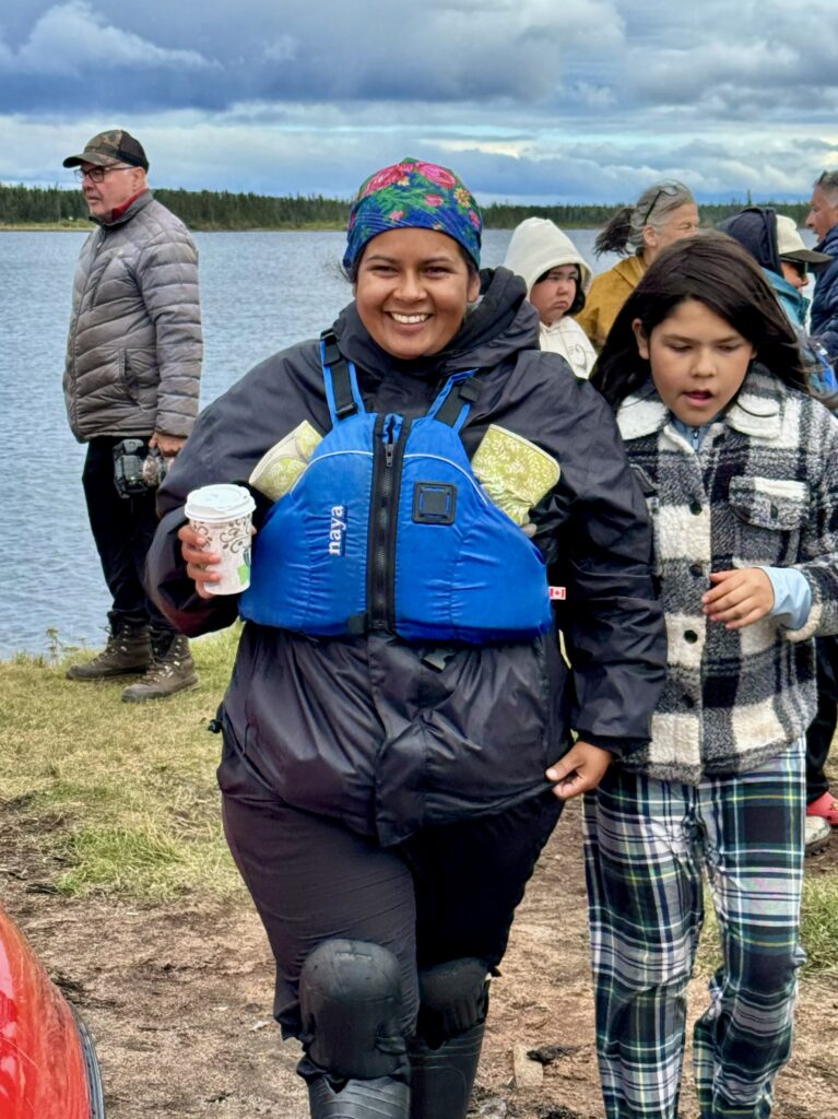 A smiling woman in a black rain jacket and blue life vest holds a cup of hot coffee while walking with a child beside her. They are part of a larger gathering by the lake, with canoes and other people in the background.