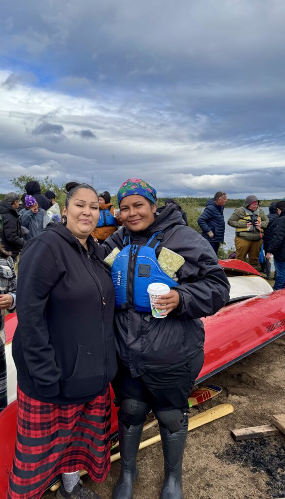 Two women standing side by side, smiling at the camera. One wears a black hoodie and red plaid skirt, while the other wears a colorful headscarf and a blue life vest, holding a cup of coffee. A red canoe and more people are visible in the background.