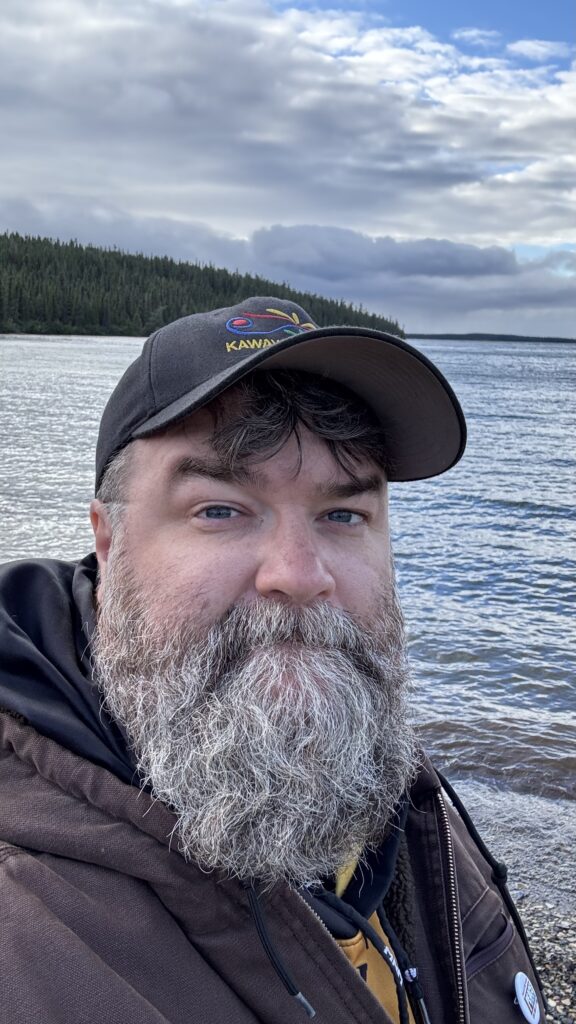 A man with a gray beard and wearing a black cap stands by a lakeshore. Behind him, the calm water and forested horizon stretch out under a partly cloudy sky, capturing the serene beauty of the natural setting.