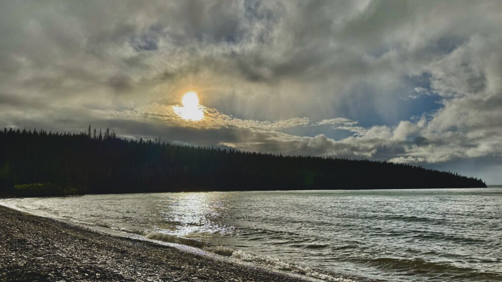 The sun breaks through clouds over a peaceful lake, reflecting on the water’s surface. The scene is framed by a dark forested shoreline, with gentle waves lapping against the rocky beach.