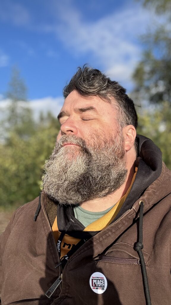 A man with a full gray beard and wearing a brown jacket stands outdoors with his eyes closed, basking in the sunlight. The blue sky and greenery in the background create a serene and peaceful atmosphere.