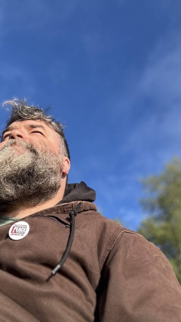 A low-angle shot of a man with a gray beard wearing a brown jacket, gazing up at the sky. The bright blue sky stretches overhead, with the edge of a few trees just visible in the background.