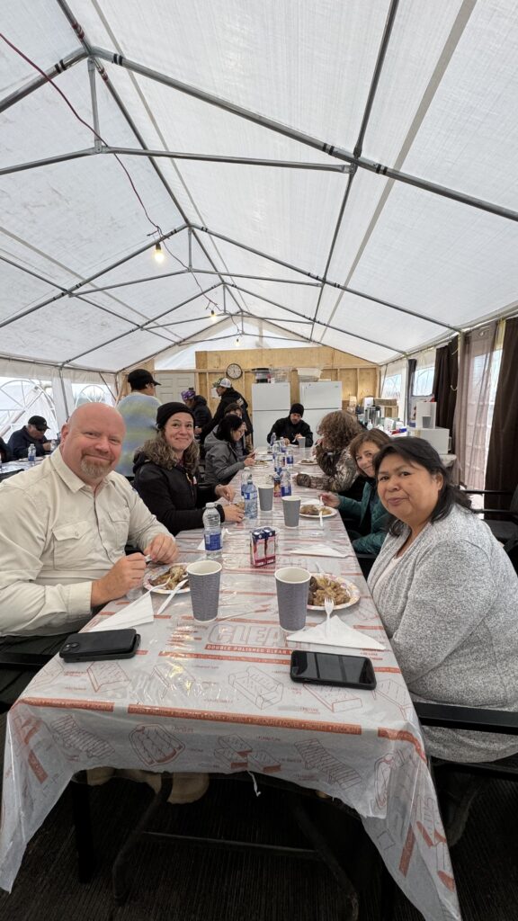 Inside a large, white, canvas dining tent, a group of people sits at a long table sharing a meal. In the foreground, Quebec's Minister of Indian Affairs, Ian Lafrenière, sits next to Chief Theresa Chemaganish. They are smiling as they enjoy dinner, surrounded by others from the camp. The table is covered with a plastic tablecloth, and plates and cups are spread out, along with some mobile phones. The atmosphere is friendly and relaxed, with people engaged in conversation.