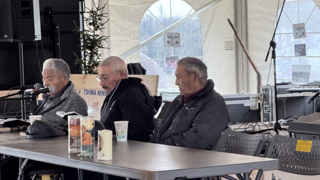 The image shows three elderly men sitting at a table during a religious service or gathering. The man on the left speaks into a microphone while the other two sit attentively beside him. The setting appears to be inside a large tent, with candles and cups placed on the table, and a mix of religious or cultural symbols displayed in the background. The atmosphere suggests a community event or worship service, where the men are likely taking part in leading or contributing to the ceremony.