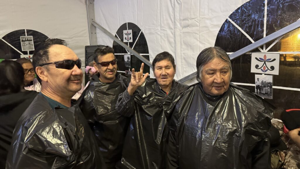 A group of four men wearing black garbage bags as makeshift rain ponchos during a talent show. Three of them are wearing sunglasses, and one is making a hand gesture. They are standing together inside a tent with photos and cultural symbols displayed on the walls behind them.