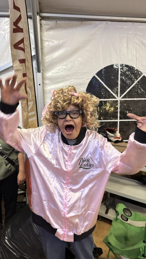 A person dressed up in a curly blonde wig, large glasses, and a shiny pink jacket with "Pink Ladies" written on the front, mimicking a character from the movie Grease. They are posing dramatically with hands up and a playful, exaggerated expression.