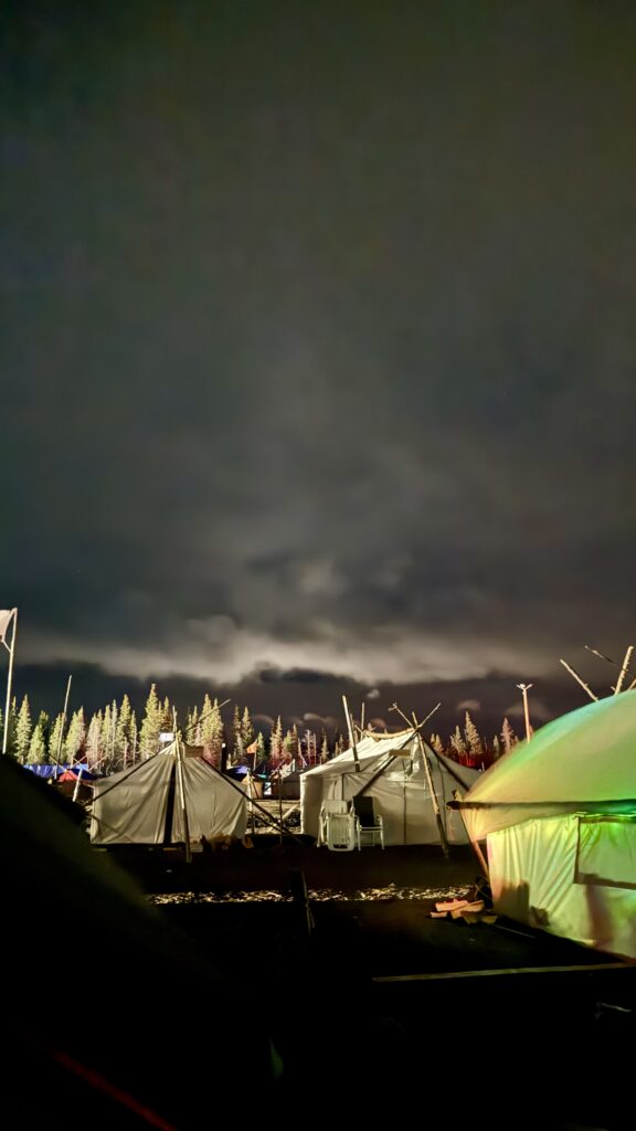 This image shows a nighttime view of a camp with several large tents illuminated by soft lighting. The sky is dark and cloudy, obscuring any chance of seeing the northern lights. In the background, a line of tall trees is faintly visible under the moody sky. The scene feels calm and quiet, as if everyone has retired for the night in preparation for the next day's events.