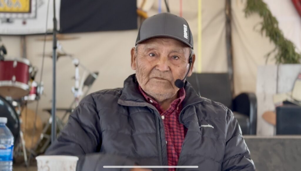 The first image shows an elderly man with a headset microphone, wearing a dark jacket and a baseball cap, seated indoors at a gathering. He looks calm and focused, possibly involved in a cultural discussion. Behind him, musical equipment like a drum set is visible.