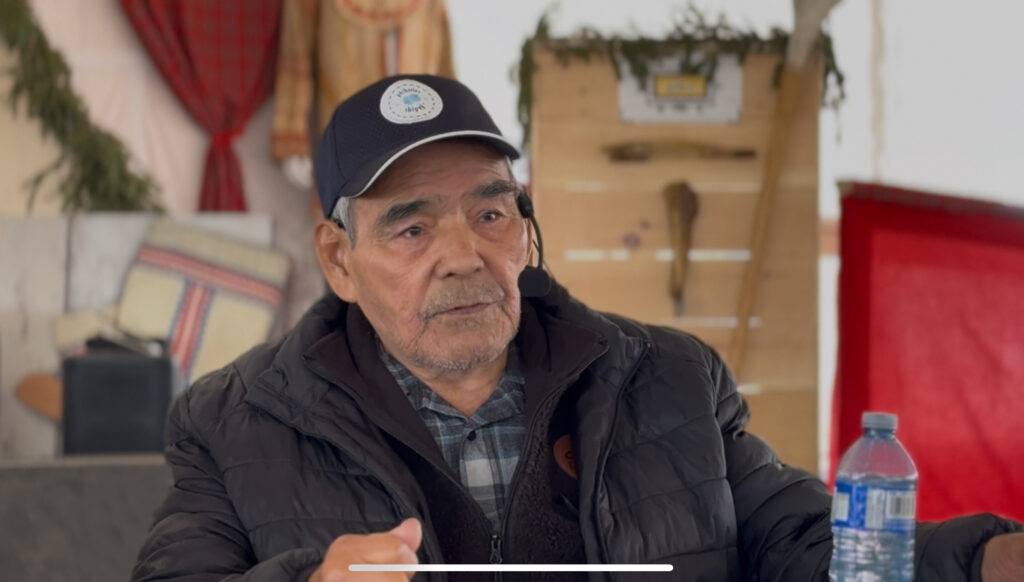 The second image features another elderly man in a dark jacket and a cap, speaking into a microphone. A water bottle sits on the table in front of him. The setting appears similar to the first image, with cultural decorations in the background, indicating a community event.