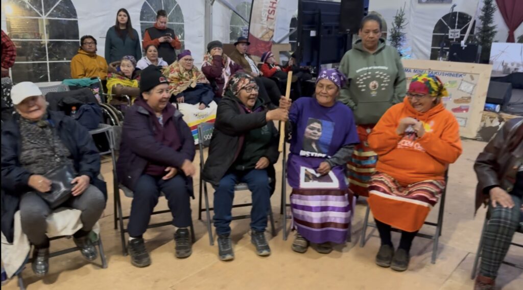 A group of elders sitting in chairs inside a tent, participating in the game "Pass The Stick." One woman, wearing a purple shirt with a printed image, is passing the stick to another participant. Everyone appears joyful, with people in the background watching and smiling. The setting is warm and communal, with colorful traditional clothing and blankets visible.