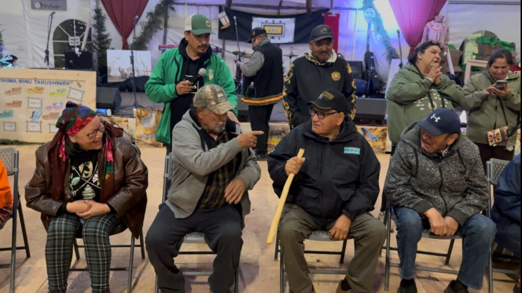 A group of elders seated in chairs, participating in "Pass The Stick." One man is pointing playfully at another who is holding the stick, while people in the background observe and laugh. The scene takes place inside a tent, with cultural decor and a stage with musical instruments visible in the background.