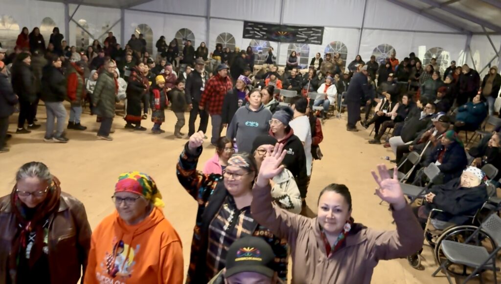 A large group of people dancing in a line inside a big tent, with smiles and raised hands. Many participants wear colorful clothing, including traditional patterns and scarves. The audience watches and cheers from the sides, creating a joyful and lively scene filled with energy and music.