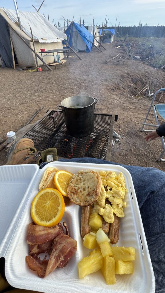 This image shows a breakfast plate resting on someone’s lap in an outdoor campsite. The plate contains bacon, orange slices, scrambled eggs, sausage, pineapple chunks, and an English muffin. In the background, tents made of tarps and canvas are set up, and a large pot is simmering over an open fire on a grill. The scene captures a peaceful moment of outdoor dining, with boots visible in the foreground and camp chairs nearby.