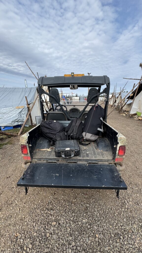 This image shows the back of a side-by-side utility vehicle with its tailgate down, loaded with bags and a hard protective case. The vehicle is parked on a gravelly area at a campsite, with tents and tarp shelters visible in the background. The sky is partly cloudy, creating a calm, transitional scene as belongings are packed up, indicating the end of the camping trip.