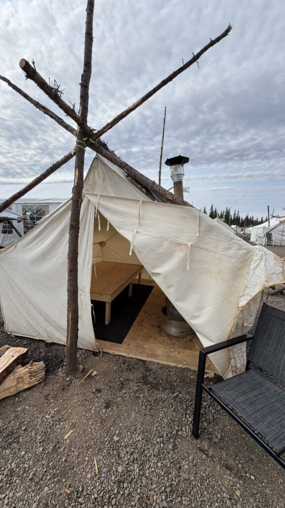 This image shows a small canvas tent with its front flaps open, revealing a simple wooden bed frame and a wood stove inside. The tent is supported by wooden poles arranged in a tripod structure, with chopped firewood stacked nearby. The sky above is partly cloudy, and surrounding tents and the natural landscape are visible in the background. The scene evokes a sense of simplicity and comfort in a rustic outdoor setting, with the tent ready for its next occupant.