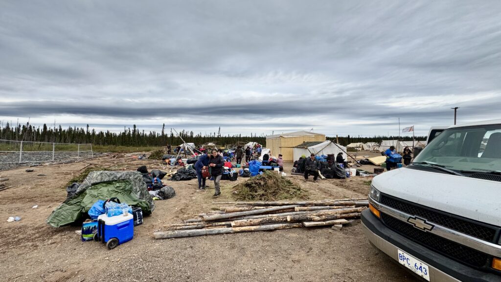 This image captures a busy campsite in the process of being packed up. Tents are being dismantled, and piles of belongings, including bags, coolers, and tarps, are scattered across the ground. People are actively moving around, organizing and packing their items. In the foreground, a white van is parked, and bundles of logs are stacked nearby. The sky is overcast, creating a somber yet transitional atmosphere as the camp prepares for departure.