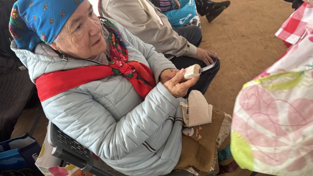 The fourth image shows an elderly woman seated, holding a small item in her hands as she participates in the gift exchange. She is wearing a light blue puffer jacket and a colorful headscarf. The atmosphere is warm and communal, with others around her engaged in the exchange event, and various bags and belongings are visible nearby.