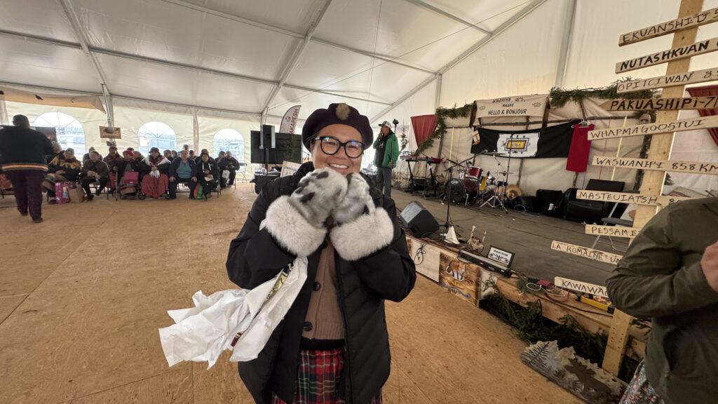 The first image shows a person smiling and holding up a pair of furry mittens, standing inside a large tent. In the background, people are seated for an event, and the stage with a microphone and musical instruments is visible.