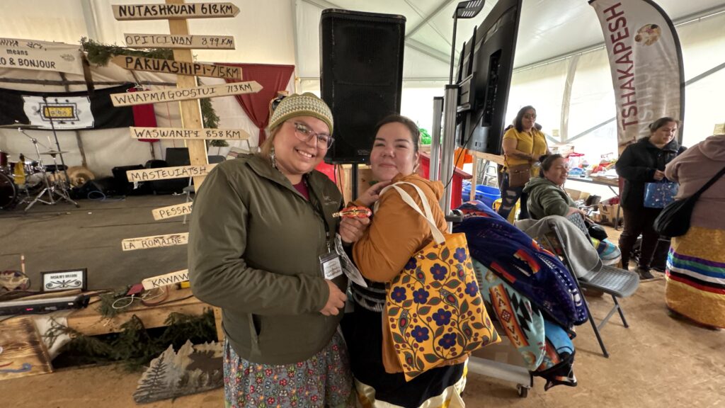 The second image features two women smiling and standing together, holding gifts, inside the large tent. Behind them, a wooden signpost lists various locations, and people can be seen chatting and exchanging gifts.