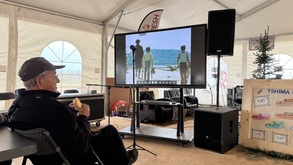 In this image, an elderly man sits in front of a TV screen, holding a piece of bread while watching a documentary. The film on screen shows three children walking along a lakeshore, and the man was one of those children in the boat featured in the film. Behind him, a large banner for "Tshima Minu" is visible along with some other decorations in the tent. The setting is intimate, capturing a moment of nostalgia.