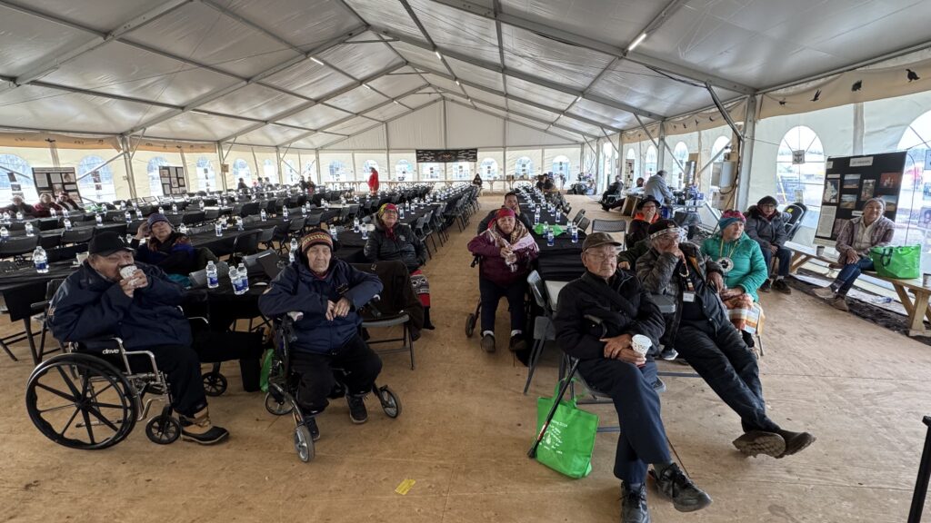 A group of elders sits in a large, mostly empty event tent, watching a presentation or documentary. Many of them are in wheelchairs or using walkers, bundled up in coats and hats, and holding drinks. The long tables in the background are set up with bottled water, but the majority of seats are unoccupied. The scene captures a moment of quiet reflection as the group takes in shared cultural traditions, emphasizing the connections between the Attikamekw and Naskapi peoples.