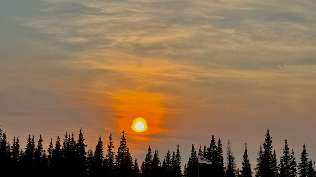 A vivid sunset fills the sky, casting a warm orange glow over the silhouetted trees. The soft, textured clouds add depth to the scene, while a single flag with a tribal emblem waves gently in the bottom right, symbolizing connection to the land and community. The peacefulness of the moment contrasts beautifully with the fiery hues of the setting sun.