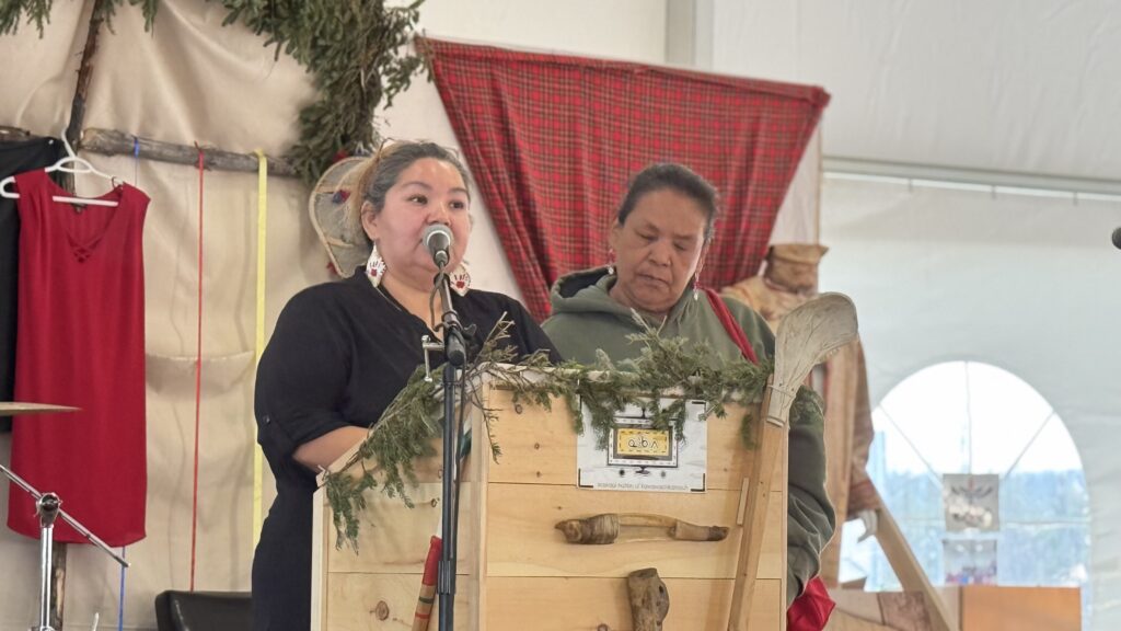 Cheyenne Peshu stands at a podium adorned with greenery, delivering a heartfelt speech, while Naomi Einish stands beside her. Both women wear traditional earrings, and their expressions convey gratitude as they thank the workers for their dedication throughout the gathering. The moment is both ceremonial and personal, especially as Cheyenne acknowledges you, calling you a "son of Kawawachikamach." The atmosphere in the tent is warm and filled with appreciation.