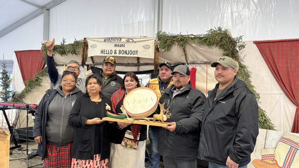 A group of people, including chiefs and representatives from various communities, stand together holding a beautifully carved gift. The gift includes a canoe, caribou, and drum, symbolizing gratitude for the gathering hosted by Kawawachikamach. A banner in the background reads 'Hello & Bonjour' in multiple languages, and festive decorations surround the scene.