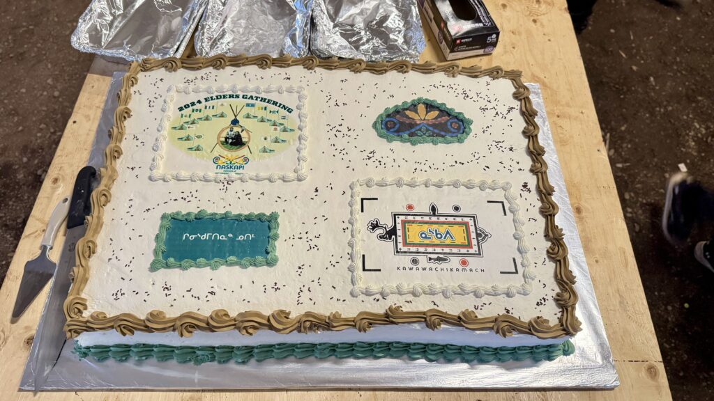 A large, celebratory cake is decorated with logos and designs representing the 2024 Elders' Gathering, featuring the event's logo, symbols of the Naskapi Nation, and the Kawawachikamach community. The cake is intricately bordered with green and tan icing, and a serving knife is placed on the wooden table beside it, ready to be shared with attendees.