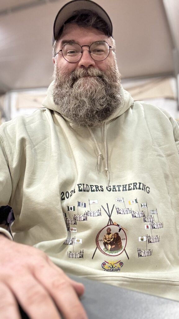 A bearded man smiling proudly while wearing a cream-colored hoodie from the 2024 Elders Gathering. The hoodie features a detailed design, including flags and a central image of an elder couple inside a teepee structure with the Naskapi logo below. This cream-colored hoodie, typically reserved for dignitaries and chiefs, was a special honor given to him at the gathering.