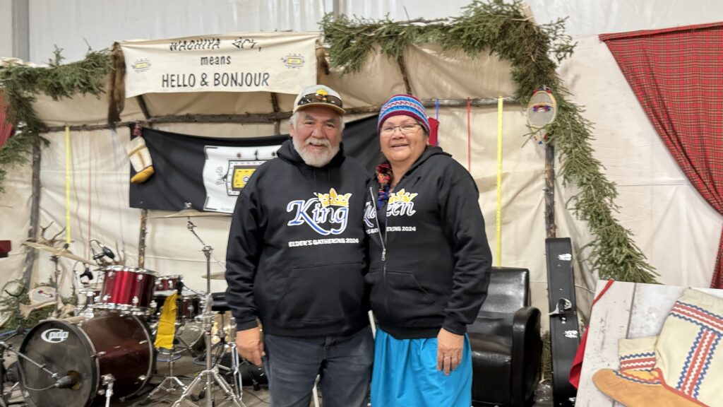 Johnny and Louisa Saganash, crowned as the king and queen of the Elders' Gathering 2024, proudly pose together in matching black hoodies. Their hoodies are printed with 'King' and 'Queen' in large font with crowns above the text, along with 'Elder’s Gathering 2024.' They stand in front of a stage decorated with evergreen branches and a banner that reads, 'Wachiya means Hello & Bonjour.'