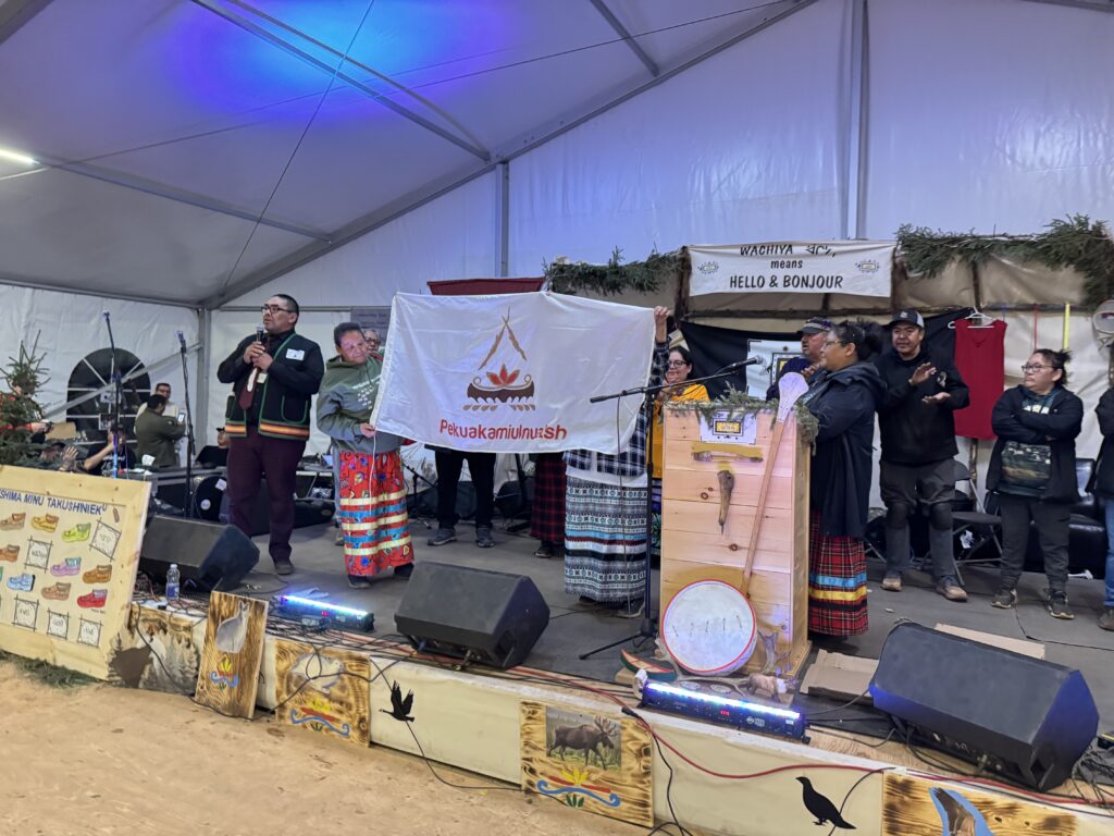 People stand on stage, holding a flag that symbolizes a village in southwest Quebec, which has been selected to host the next Elders Gathering. A speaker stands at the podium, while others in traditional attire and audience members listen and observe the announcement.