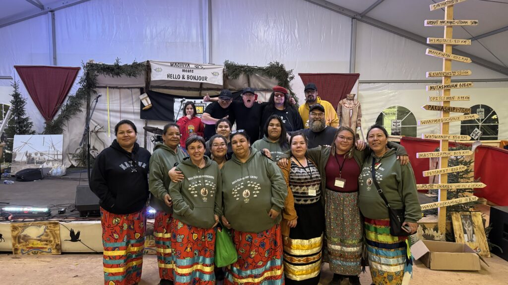 A large group photo of the event crew standing proudly together on stage after the gathering. The women in the front row wear colorful, traditional skirts, while others in the group are dressed in event hoodies and casual wear. Everyone is smiling, celebrating their hard work and successful collaboration, with event decorations and directional signs in the background.